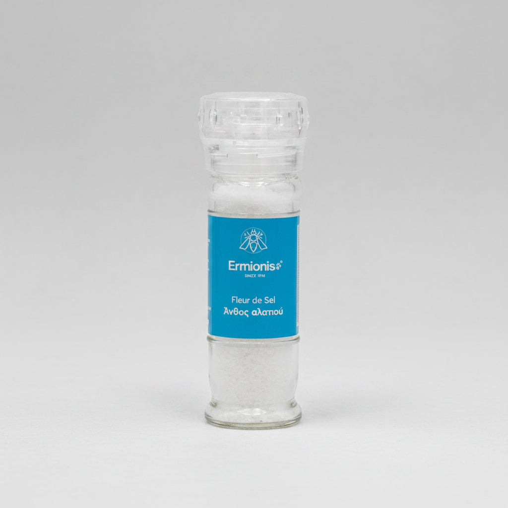 A clear glass salt shaker filled with white salt, featuring a blue label with the brand name Ermionis Greek fleur de sel sea salt mill, 75g