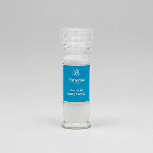 A clear glass salt shaker filled with white salt, featuring a blue label with the brand name Ermionis Greek fleur de sel sea salt mill, 75g