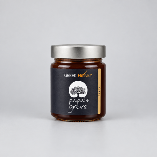 A glass jar of Greek Honey from Papa's Grove, filled with dark amber honey and featuring a black label with a white tree logo and a silver metal lid, set against a neutral gray background.