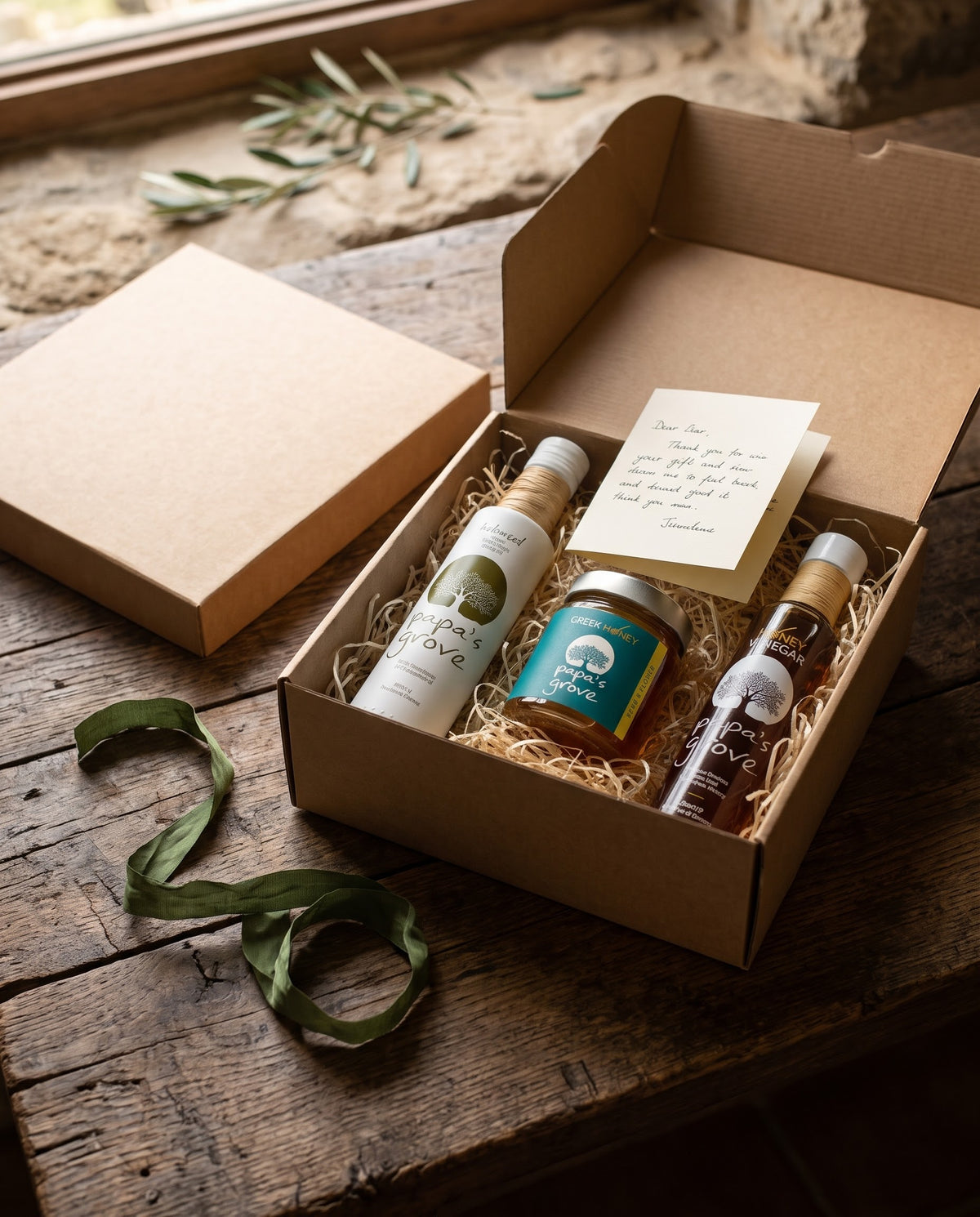 Papa's Grove extra virgin olive oil, honey, and honey vinegar styled in a craft giftbox, with t handwritten card on a wooden table in a rustic setting.