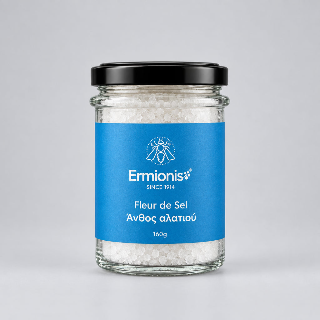 A glass jar of Ermionis Fleur de Sel sea salt with a black lid and a blue label, in a light grey backdrop