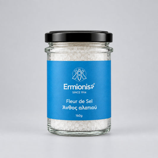 A glass jar of Ermionis Fleur de Sel sea salt with a black lid and a blue label, in a light grey backdrop