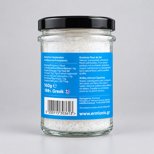A glass jar of Greek Ermionis salt flakes with a black lid and a blue label, with a light grey background.