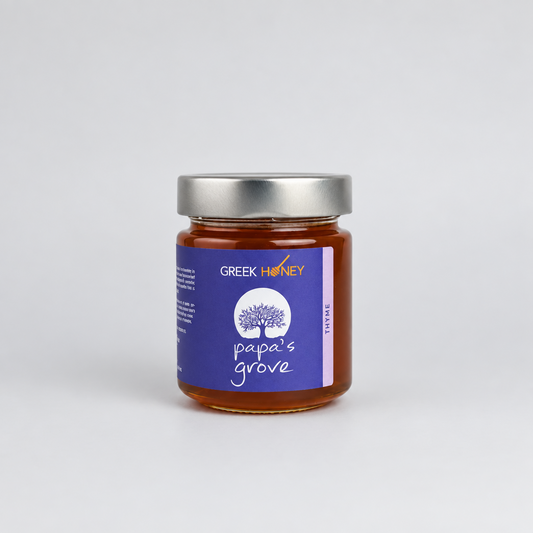 A glass jar of Greek Honey from Papa's Grove, filled with amber honey and featuring a purple label with a white tree logo and a silver metal lid, set against a light gray background.